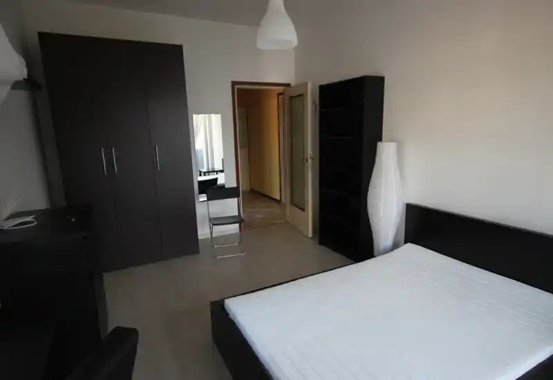 Camera da letto minimalista con letto matrimoniale, mobili in legno scuro e una lampada da terra alta. Pareti neutre e armadio coordinato creano un'area notte pulita e moderna.