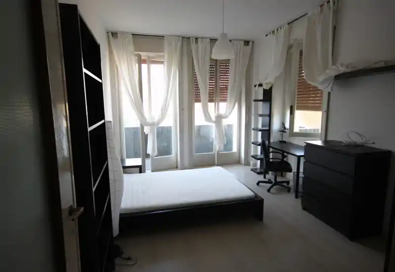 Camera da letto luminosa e minimal con un letto basso a piattaforma davanti a tre finestre alte con tende bianche leggere; scrivania e mobili scuri completano l’arredo pulito e arioso.