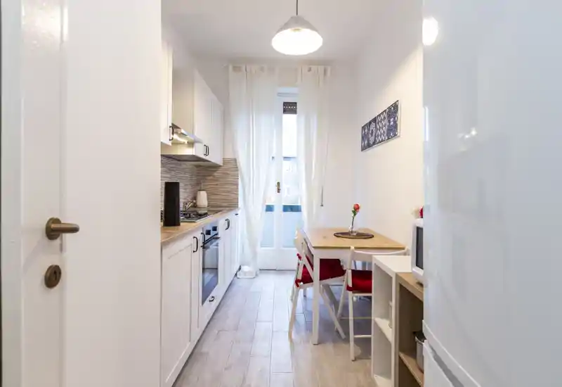 Cucina compatta e luminosa con pensili bianchi, forno incassato e un piccolo tavolo da pranzo vicino alla porta finestra — perfetta per pasti accoglienti.