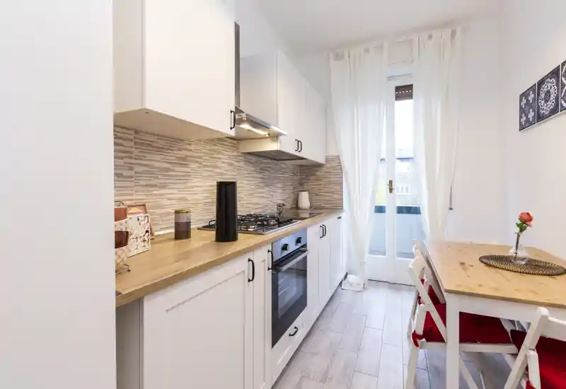 Bright, modern galley kitchen with white cabinetry, wooden countertops, built-in oven and a small dining table by a glass door letting in natural light.