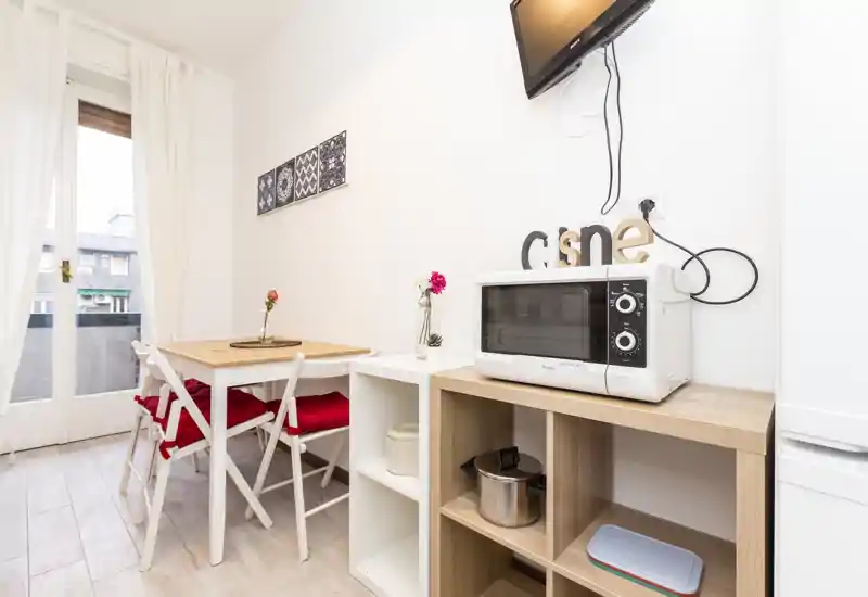Small bright kitchen corner with a compact dining table, chairs with red cushions, a microwave on open shelving and a wall-mounted TV; clean, well-lit and neatly styled for short-stay rentals.