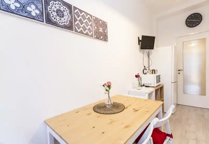 Small dining area with a wooden table for two, simple decor and a vase with flowers; compact kitchenette elements (microwave, fridge) visible in the background.