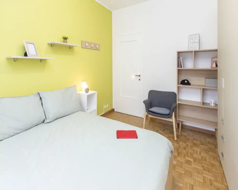 Bright, modern bedroom with a large bed dressed in pale linens, a yellow accent wall, bedside shelving and a cozy armchair — well lit and simply styled for rental listings.