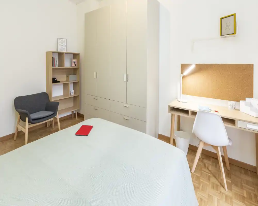 Bright, minimalist bedroom with a neatly made bed in the foreground, a wardrobe and a study desk with cork board. Natural light and simple Scandinavian-style furniture create a calm, welcoming atmosphere.