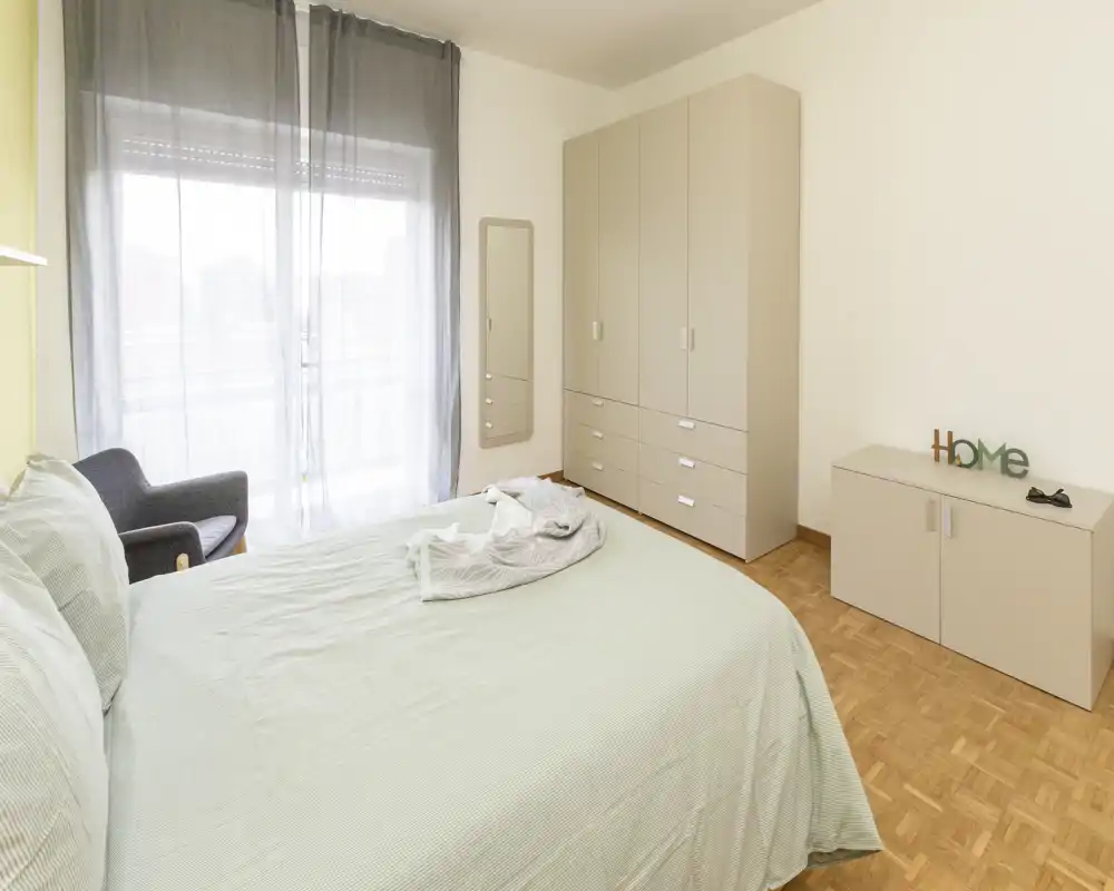 Chambre lumineuse au style simple avec un grand lit, une armoire et une baie vitrée donnant sur le balcon. Décoration minimale et textiles doux pour une ambiance calme, parfaite pour une annonce.