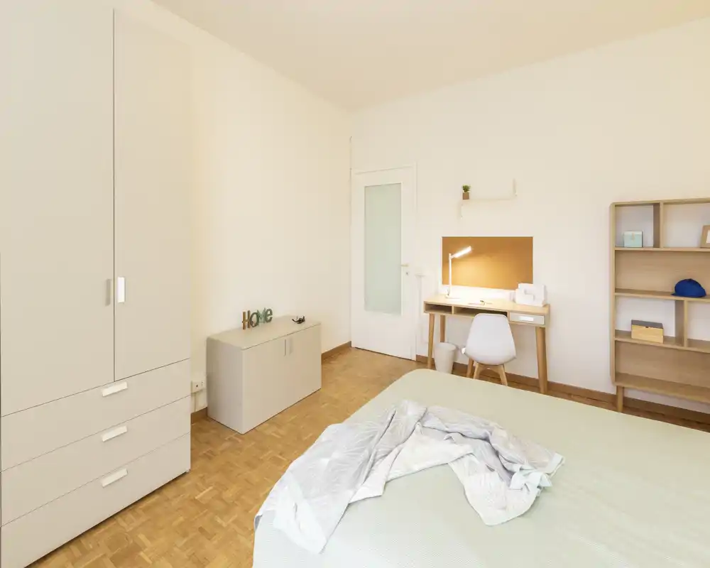 Chambre lumineuse et minimaliste avec lit au premier plan, petit bureau avec lampe, armoire et étagère ; tons neutres et parquet en bois créent une atmosphère apaisante.