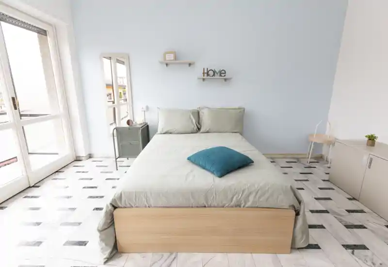 Chambre lumineuse et minimaliste avec lit double centré, mur bleu pâle et grande fenêtre apportant une belle lumière naturelle — idéale pour les annonces.