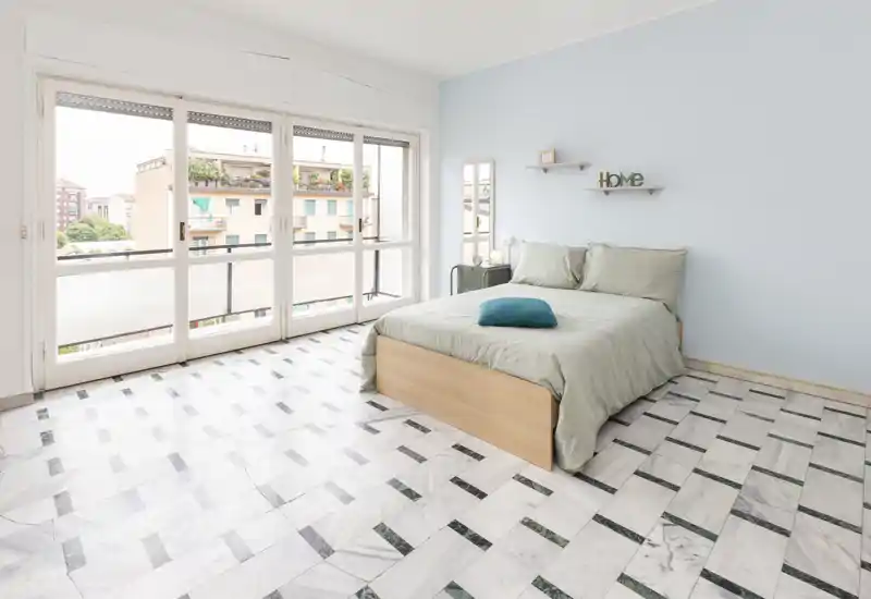 Chambre lumineuse et minimaliste avec un lit double près de grandes baies vitrées ; murs pastel et sol effet marbre pour une ambiance moderne et aérée.
