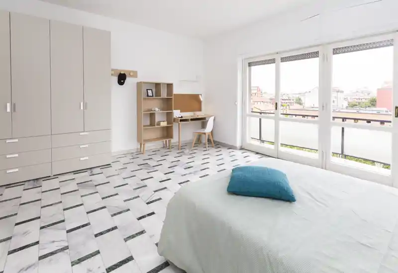 Chambre lumineuse et moderne avec un lit bien fait, une grande armoire et un petit bureau près de larges fenêtres donnant sur le balcon — ambiance aérée et design minimal, parfaite pour un séjour confortable.