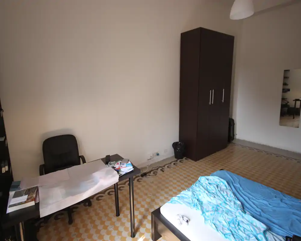 Chambre simple et lumineuse avec un lit double, une armoire et un bureau ; le carrelage à motif apporte du caractère et la lumière naturelle éclaire la pièce.