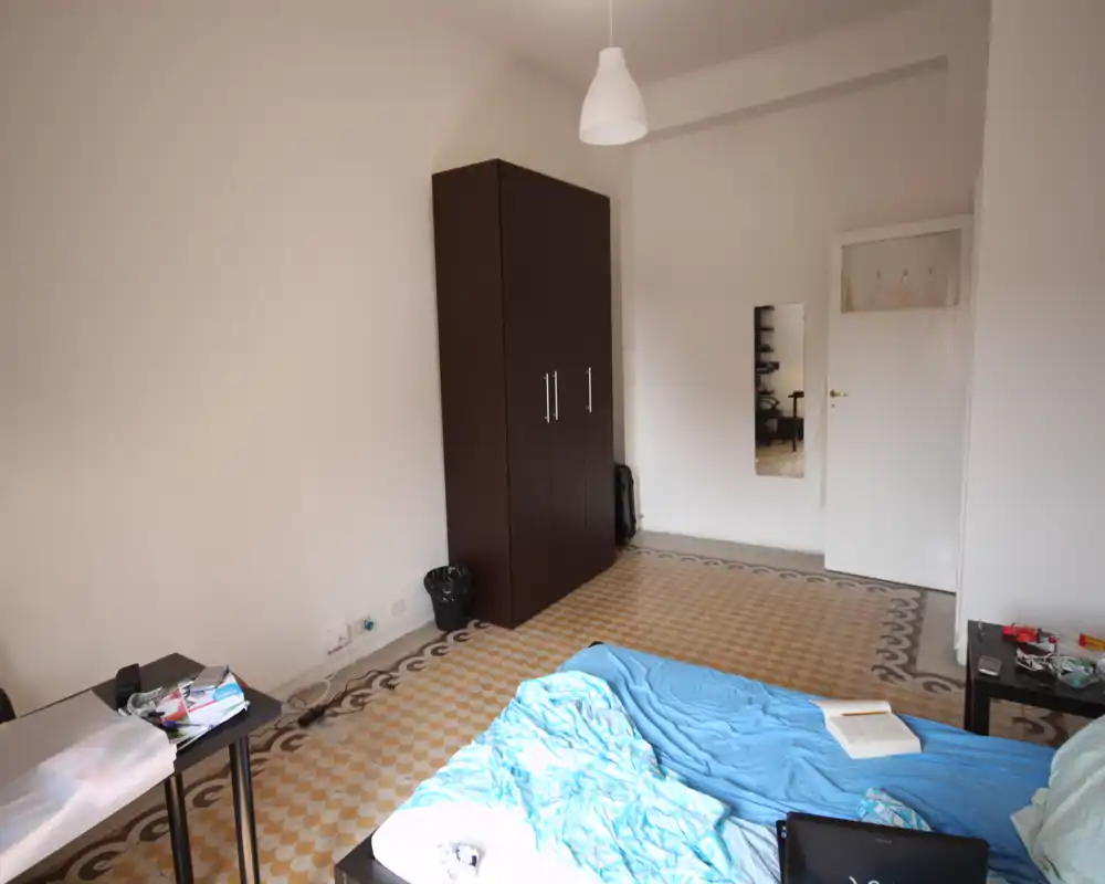Chambre simple avec un lit double au premier plan, couette bleue en désordre, grande armoire sombre, sol carrelé à motifs et miroir sur le mur du fond. Pièce bien éclairée et habitée, adaptée à une annonce mettant en valeur le sol et les rangements.