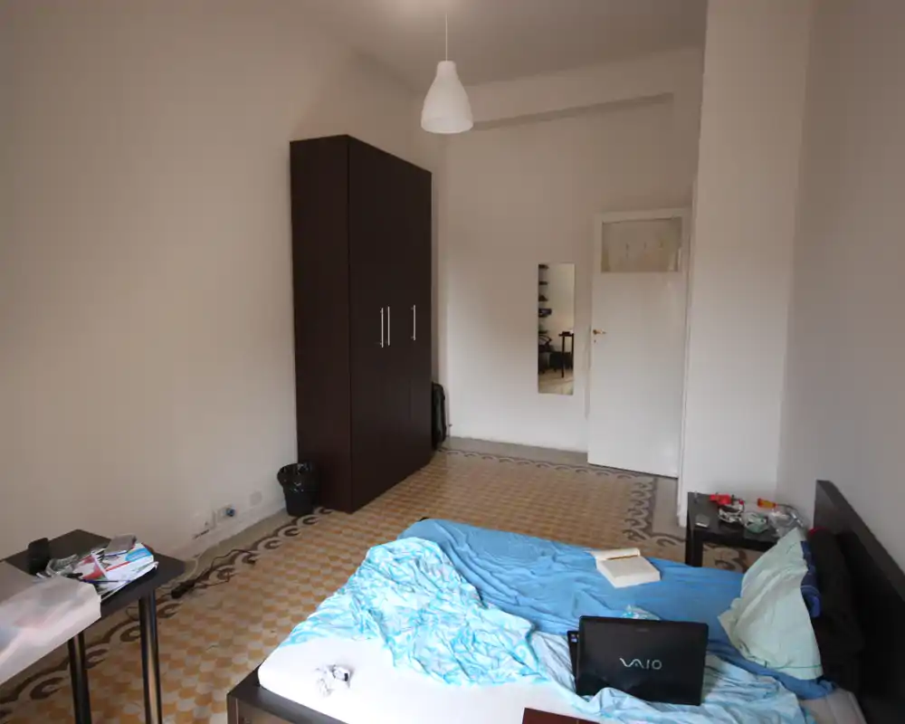 Chambre lumineuse et sobre avec lit double, armoire et sol carrelé à motifs ; quelques objets personnels sur la table de chevet et le bureau donnent un aspect habité.