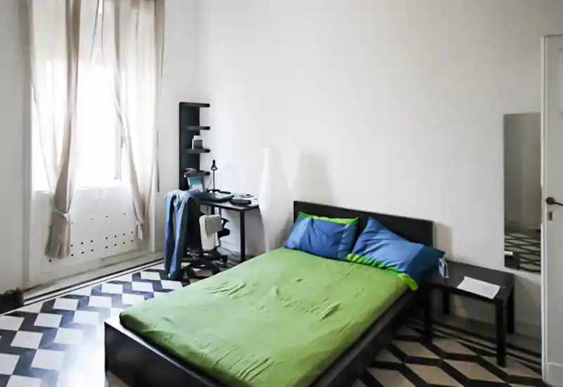 Chambre lumineuse et minimaliste avec un lit double, une literie colorée et un petit bureau près de la fenêtre. Le sol carrelé à motifs et les rideaux hauts créent une atmosphère élégante et aérée, idéale pour la location courte durée.