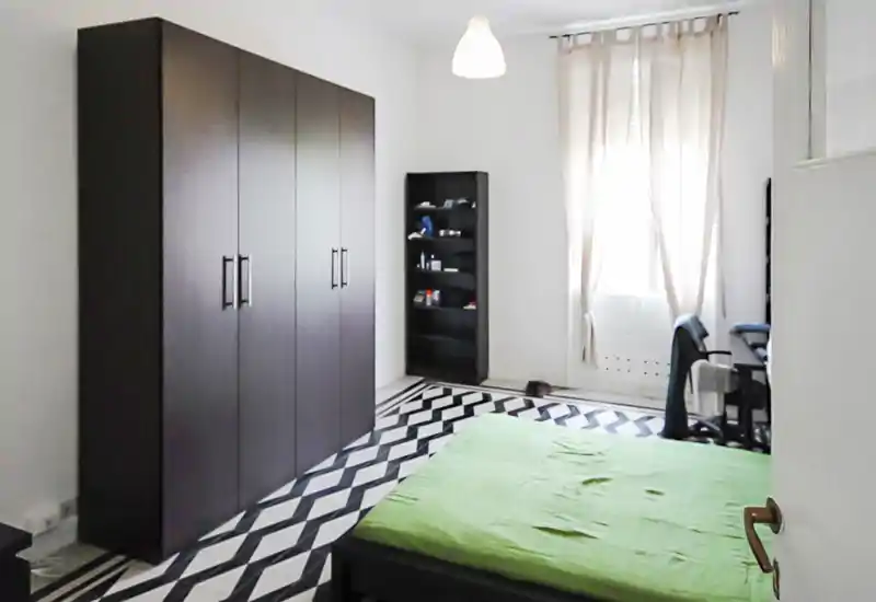 Chambre lumineuse et sobre avec lit double, grand placard et sol à motifs géométriques. Grande fenêtre apportant une belle lumière naturelle et un coin bureau fonctionnel.
