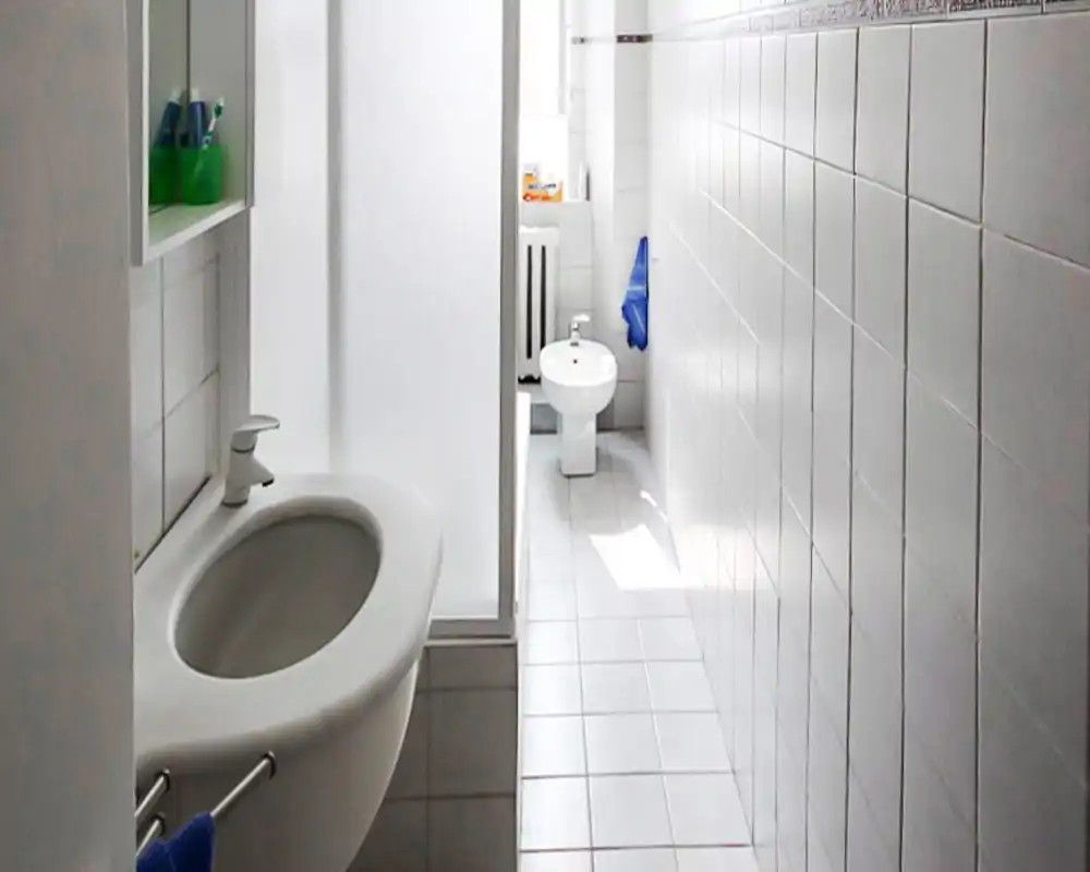 Salle de bain étroite et lumineuse avec carrelage blanc, lavabo mural et bidet au fond. Agencement simple et propre, éclairé par une petite fenêtre.
