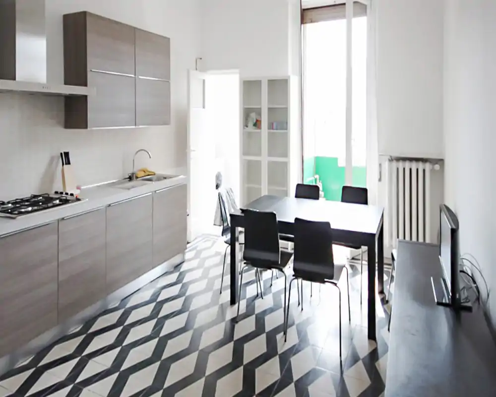 Cuisine ouverte lumineuse avec meubles effet bois, plaque gaz et table à manger pour quatre près d'une grande fenêtre. Le sol en carrelage à motif et le mobilier minimaliste dégagent une ambiance moderne et aérée.