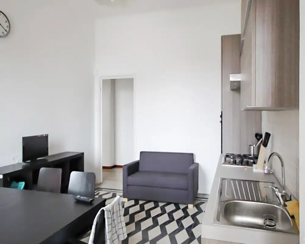 Espace de vie en open-space avec kitchenette : petit canapé, table à manger et plan de cuisson avec évier visibles. Lignes épurées et sol à motifs apportant du caractère.