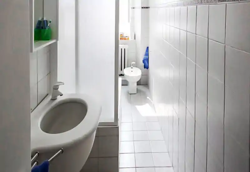 Salle de bains étroite et lumineuse avec murs carrelés blancs, petit lavabo mural et bidet au fond ; installations simples et lumière naturelle par une petite fenêtre.