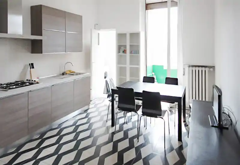 Cucina moderna e luminosa con pensili grigi, piano cottura integrato, lavello e un tavolo da pranzo nero con sedie. Ampia finestra che offre luce naturale e pavimento a motivi che dona carattere.
