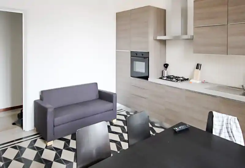 Espace de vie open‑plan avec un petit canapé gris et une cuisine intégrée aux façades effet bois et une plaque gaz. Le carrelage géométrique et la table noire donnent une atmosphère moderne et cohérente, idéale pour un petit appartement.