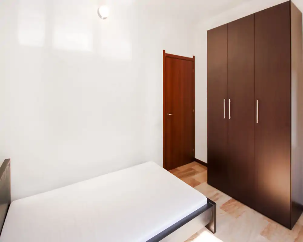 Simple, bright bedroom with a visible double bed frame and a large dark-wood wardrobe. Neutral walls and parquet flooring create a clean, minimal look ideal for short-term rentals.