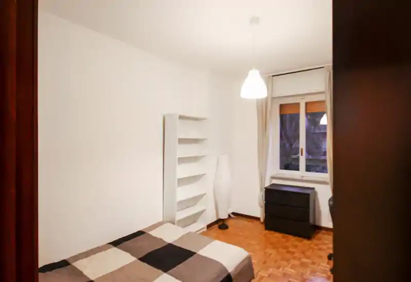 Dormitorio luminoso y con mobiliario sencillo: cama doble, estantería alta, lámpara de pie y ventana que aporta luz natural. Suelo de parquet cálido y decoración neutra, ideal para anuncios de alquiler.