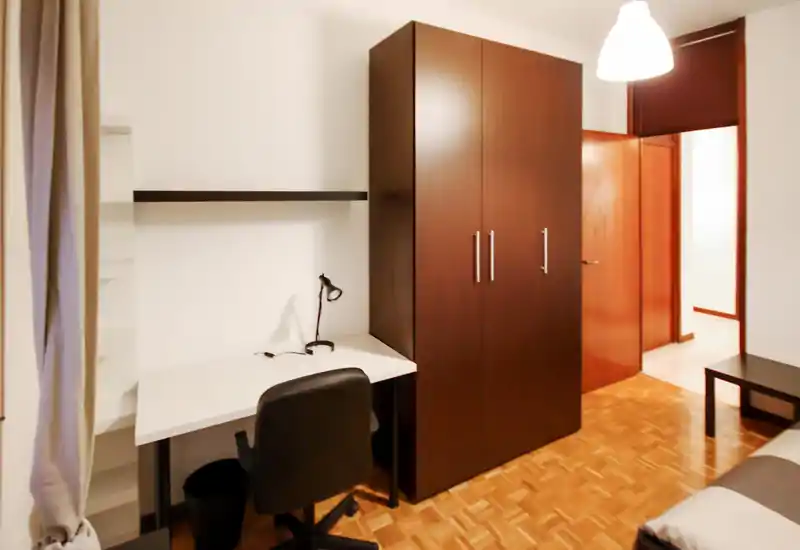 Dormitorio compacto con armario de madera oscura, un pequeño escritorio blanco y una lámpara de trabajo, y un ligero vistazo de la cama a la derecha. El suelo de parquet y la iluminación cálida aportan un ambiente limpio y funcional, ideal para estudiar o estancias cortas.