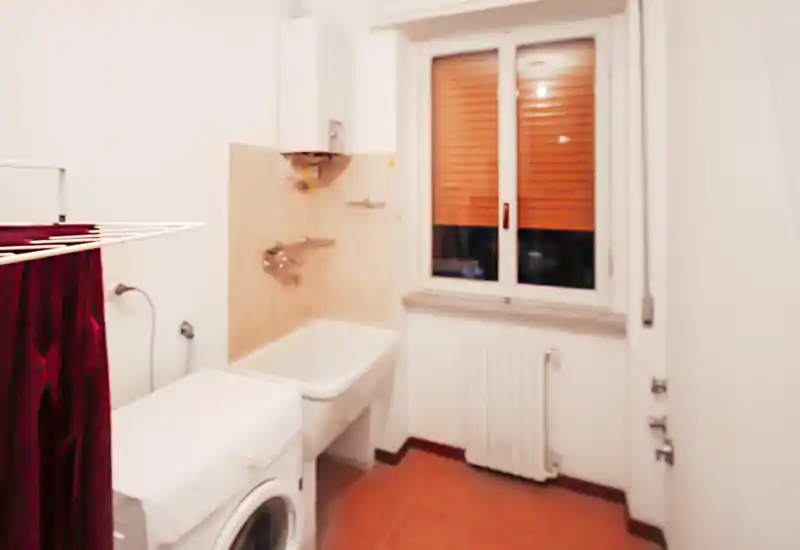 Baño / lavandería compacta con lavadora, caldera mural, pila y salpicadero de azulejos junto a la ventana — distribución práctica para lavado y limpieza.