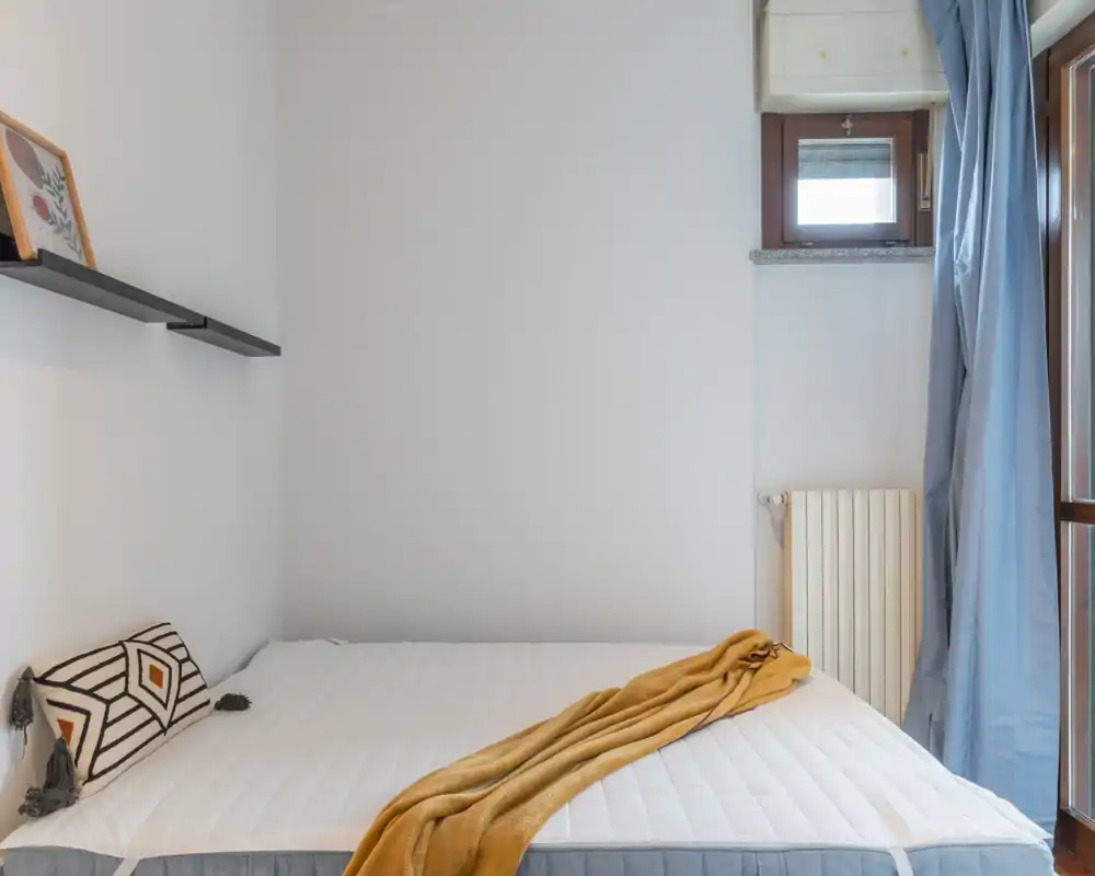 Bright, minimalist bedroom with a large bed dressed in white linens, a mustard throw and a decorative pillow; natural light enters through the window and blue curtains.