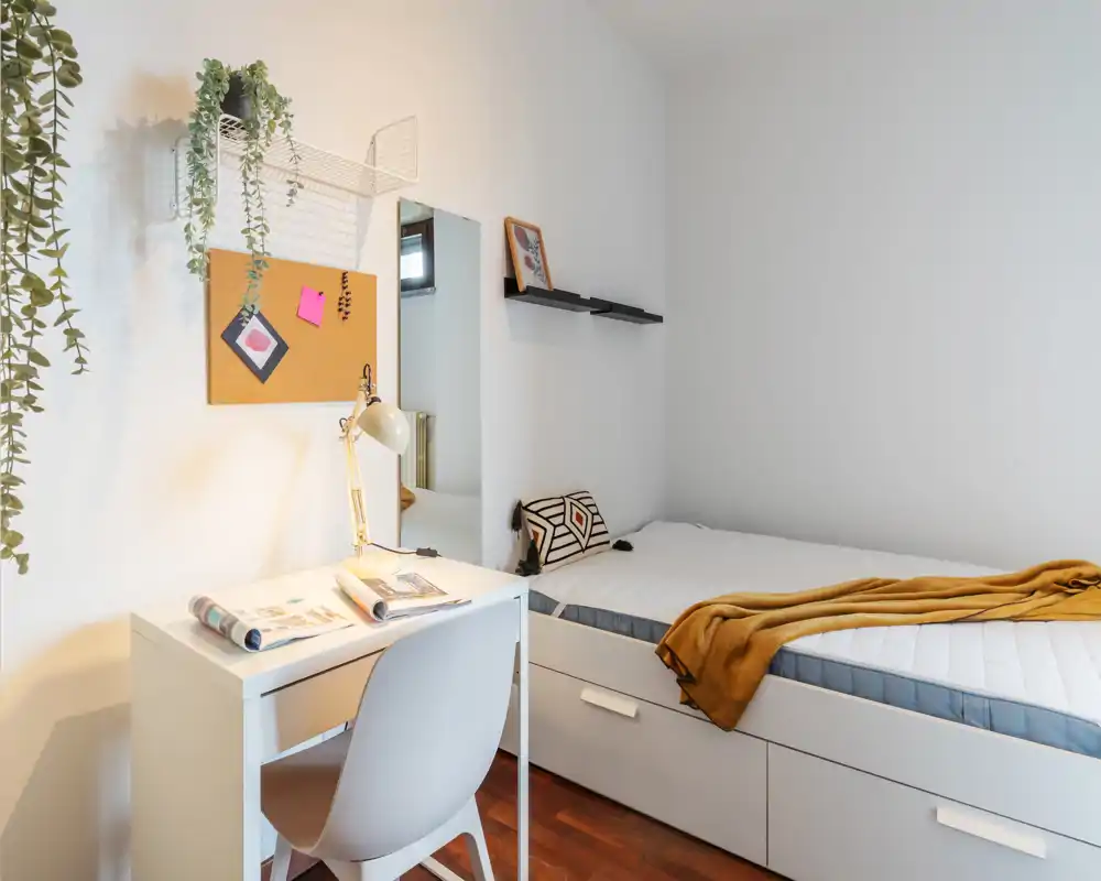 Bright, neatly styled single bedroom with a white desk, comfortable single bed with storage drawers and warm accents like a mustard throw and decorative pillow.