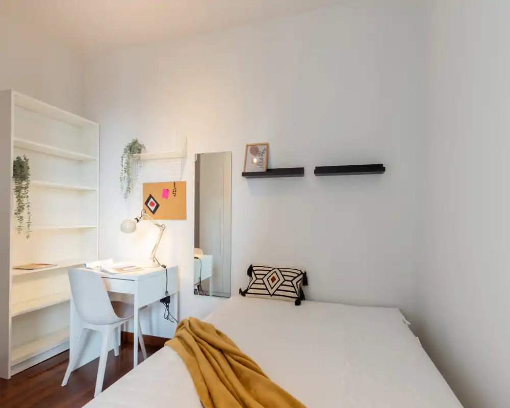 Bright, minimal bedroom with a neatly made bed, decorative pillow and a mustard throw; a small desk and shelving create a functional workspace.