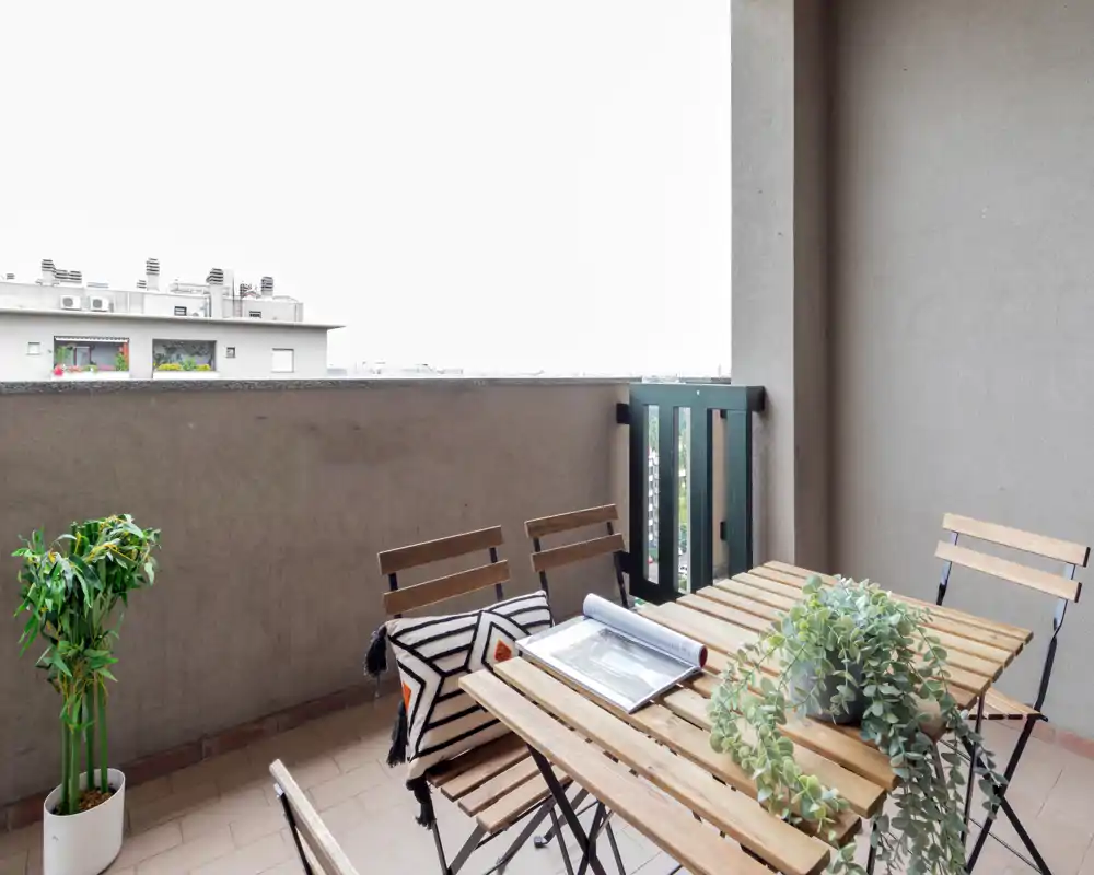Sunny balcony with a wooden folding table and chairs, potted plants and a decorative cushion — a cozy outdoor spot for dining or reading.
