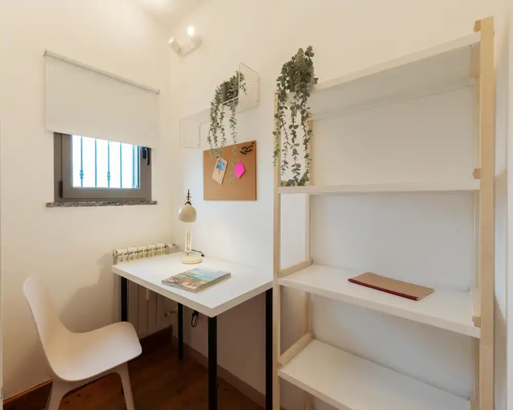 Angolo lavoro compatto e ordinato con scrivania bianca, sedia, scaffale e piante ornamentali sospese; luce naturale e lampada calda creano un'atmosfera tranquilla.