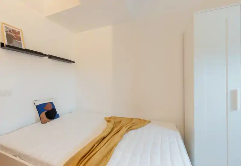 Camera da letto luminosa e minimal con armadio bianco e letto matrimoniale ben rifatto, valorizzato da un cuscino decorativo e una coperta color senape.