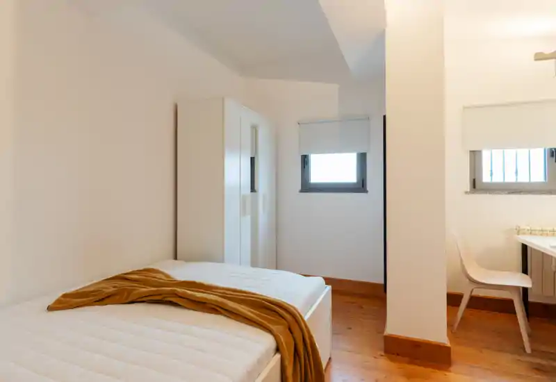 Camera da letto luminosa e minimal con letto comodo, pavimento in legno, armadio bianco e un caldo plaid ocra; è presente anche una piccola scrivania vicino alla finestra.