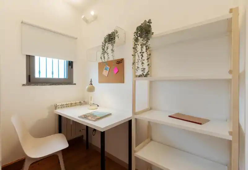 Angolo lavoro compatto e ordinato con scrivania bianca, sedia, scaffale e piante ornamentali sospese; luce naturale e lampada calda creano un'atmosfera tranquilla.