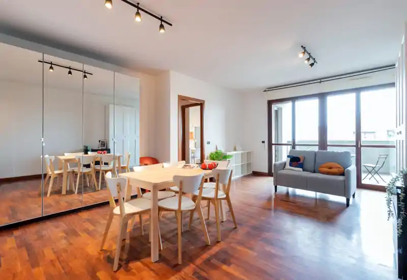 Ampio soggiorno moderno con pavimento in legno, tavolo da pranzo e un divano due posti accogliente vicino a grandi finestre che si affacciano sul balcone.