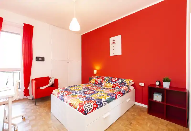 Camera da letto luminosa e ben arredata con letto contenitore bianco, biancheria floreale colorata e parete d'accento rossa. La stanza presenta una lampada da comodino, un piccolo scaffale e una poltrona rossa vicino alla finestra, per un ambiente accogliente e moderno.