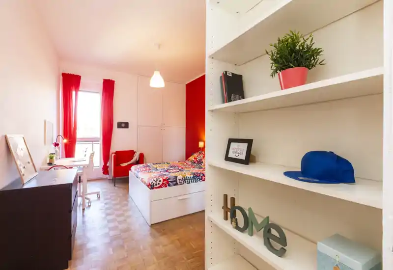 Camera da letto luminosa e colorata con letto singolo, armadio a muro e scrivania vicino alla finestra. Tende rosse vivaci e biancheria a pattern donano personalità, mentre la libreria in primo piano offre spazio e profondità.