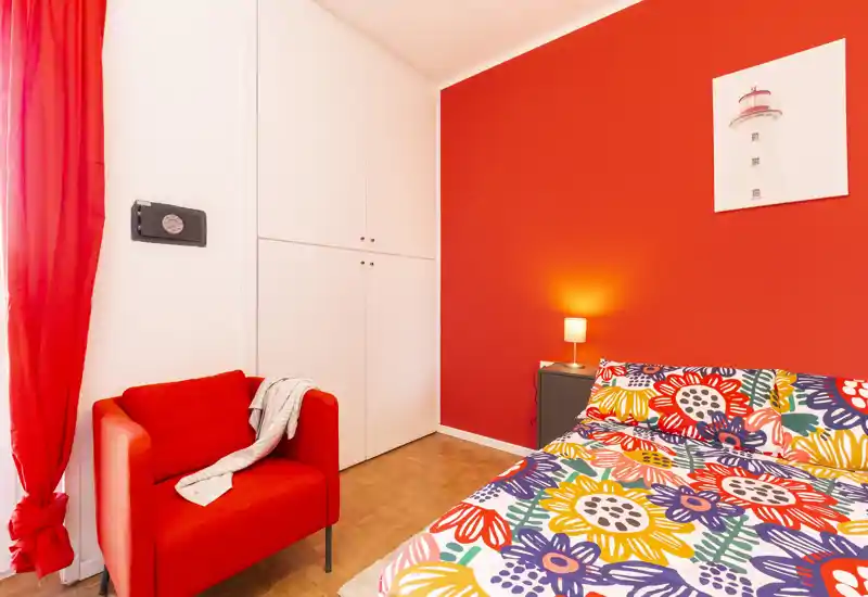 Camera da letto luminosa e vivace con letto a fantasia, lampada da comodino e una confortevole poltrona rossa accanto a un armadio a muro bianco.