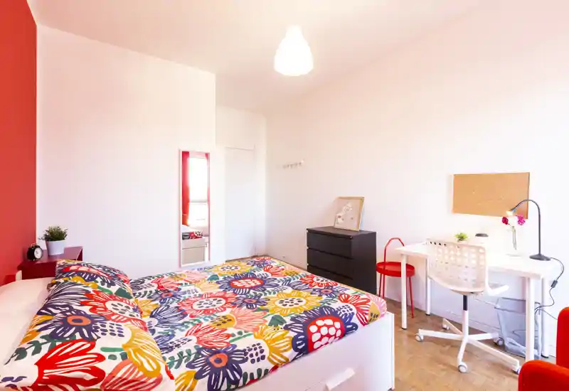 Camera da letto moderna e luminosa con una copertura del letto colorata, pareti bianche, scrivania e comò: ideale per uno studente o una persona che cerca un ambiente vivace.