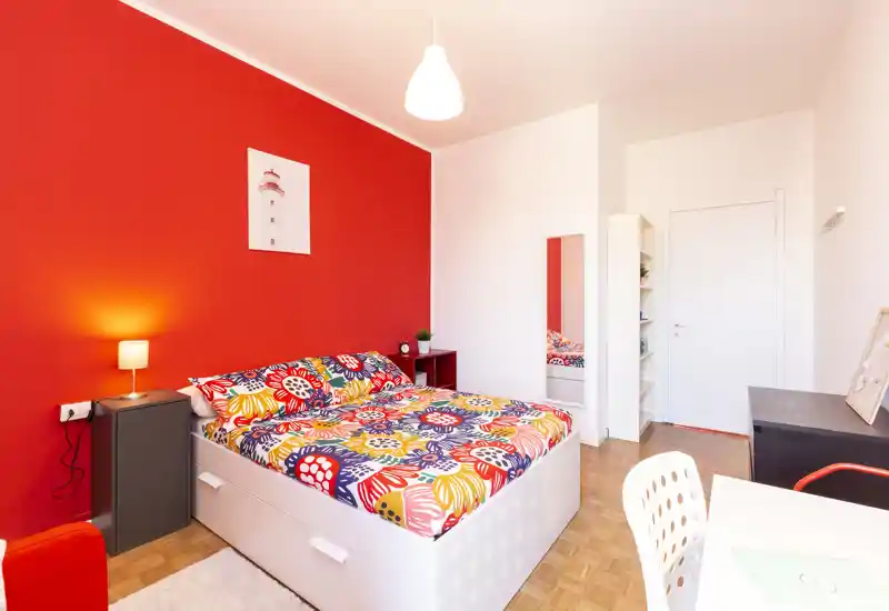 Camera da letto luminosa e ben arredata con un grande letto matrimoniale colorato contro una parete rossa a effetto, lampada da comodino e zona scrivania — perfetta per un affitto moderno e vivace.