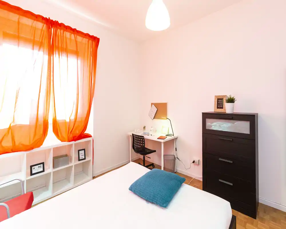 Dormitorio luminoso y bien presentado con cama visible, cojín turquesa, cómoda negra y un pequeño escritorio junto a la ventana con cortinas naranjas que dejan pasar la luz.