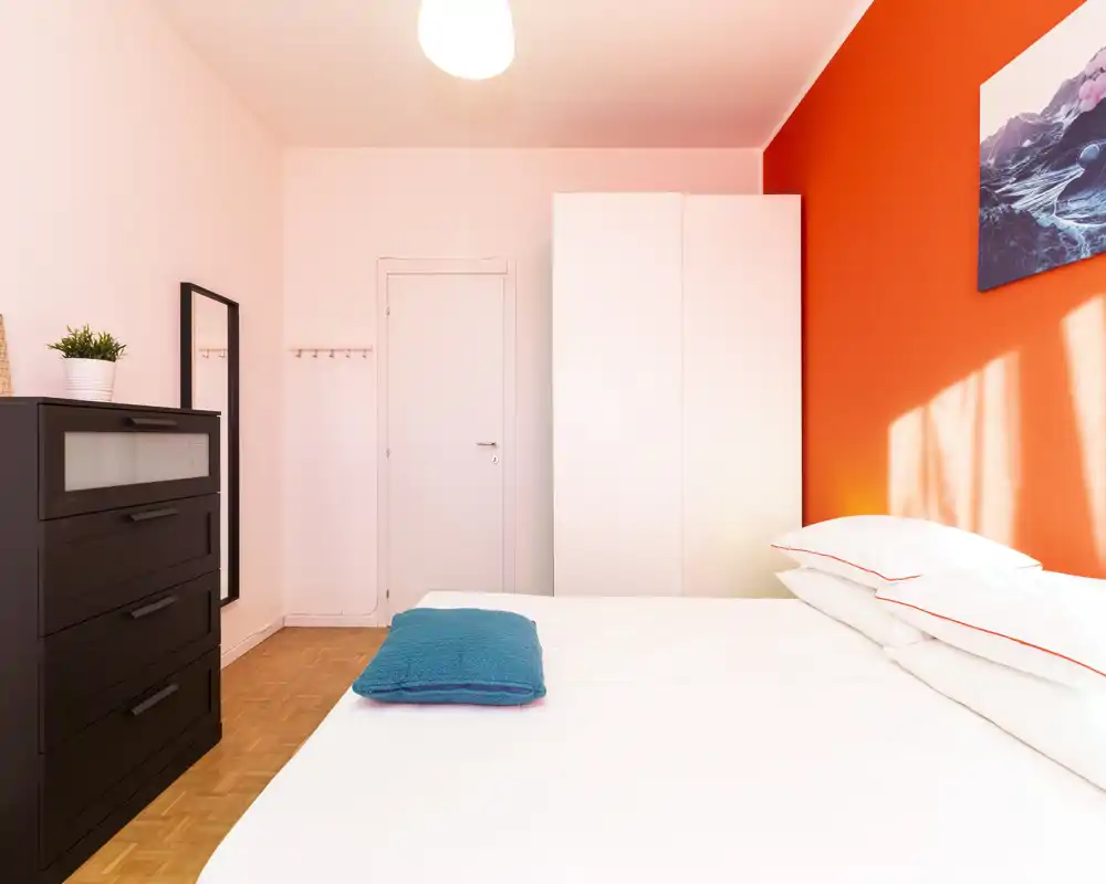 Dormitorio moderno y luminoso con una gran cama blanca en primer plano, pared de acento naranja, armario y cómoda — bien iluminado y acogedor.
