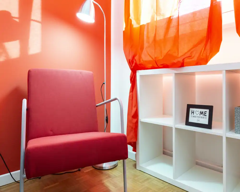 Rincón acogedor con sillón rojo, lámpara de pie y estantería bajo una cortina luminosa: un pequeño salón/rincón de lectura con tonos cálidos.