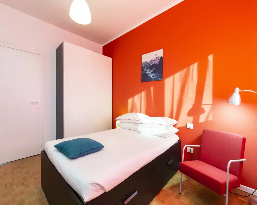 Dormitorio individual moderno y luminoso con cama bien hecha, pared naranja llamativa y un sillón cómodo — ideal para estancias cortas.