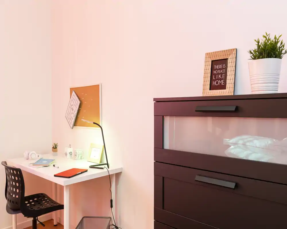 Rincón de dormitorio con cómoda oscura que muestra almohadas tras un panel de vidrio, un escritorio blanco brillante con lámpara y silla, y detalles decorativos mínimos (planta y marco).