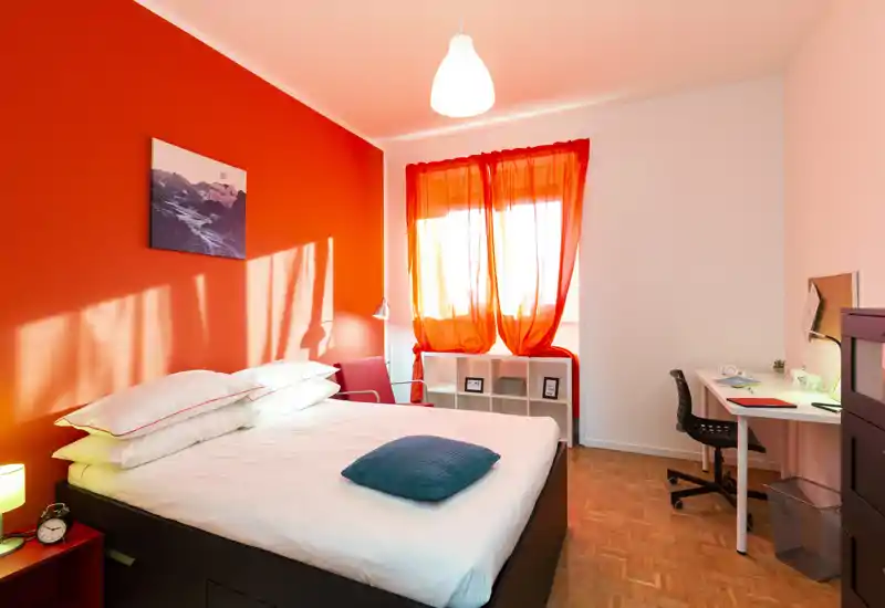 Dormitorio luminoso y con estilo, con cama doble centrada, pared roja de acento y cortinas translúcidas que dejan entrar luz natural. Un escritorio y estanterías ofrecen un rincón de trabajo funcional.