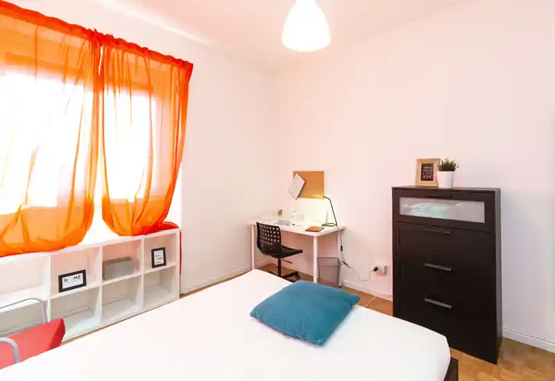 Camera da letto luminosa e ben allestita con letto visibile, cuscino turchese, comò nero e una piccola scrivania vicino alla finestra con tende arancioni che filtrano la luce.
