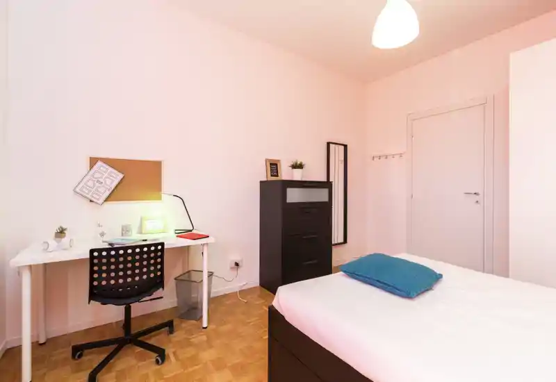 Dormitorio luminoso y minimalista con cama doble, escritorio y cómoda. Suelo de parquet y decoración sencilla que crean un espacio ordenado y funcional, ideal para estudiantes o profesionales.